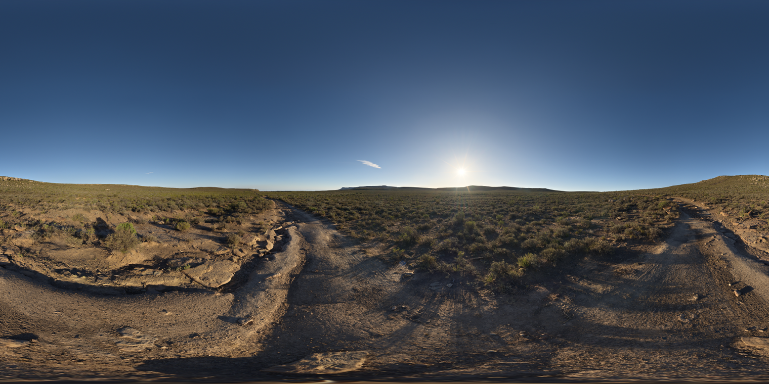 Unclipped, free 24K HDRI: sun low over a scrubby dirt road beneath a clear blue sky, warm golden-hour light casting long shadows with crisp contrast.