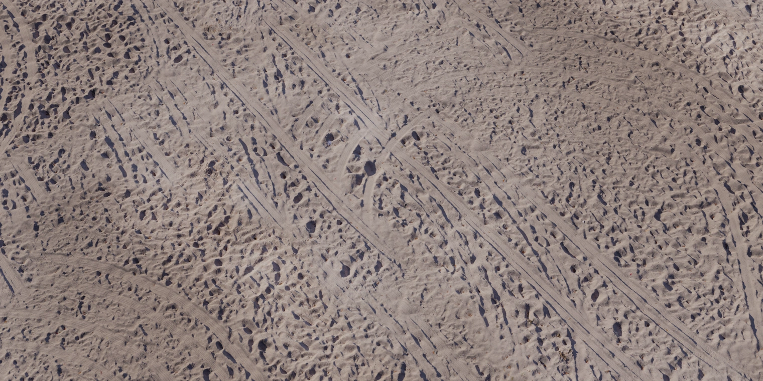 Free 8K texture of aerial coastal sand: tan, compacted ripples with tire and foot ruts, fine grain, rough bumps and subtle wet-dark patches.