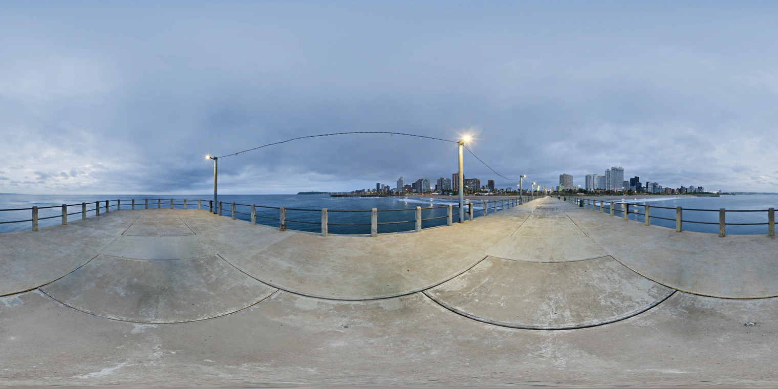Free 24K unclipped HDRI of an urban pier at dawn. Cool blue hour sky, soft overcast light, calm sea, with warm streetlights and pale sunrise glow.
