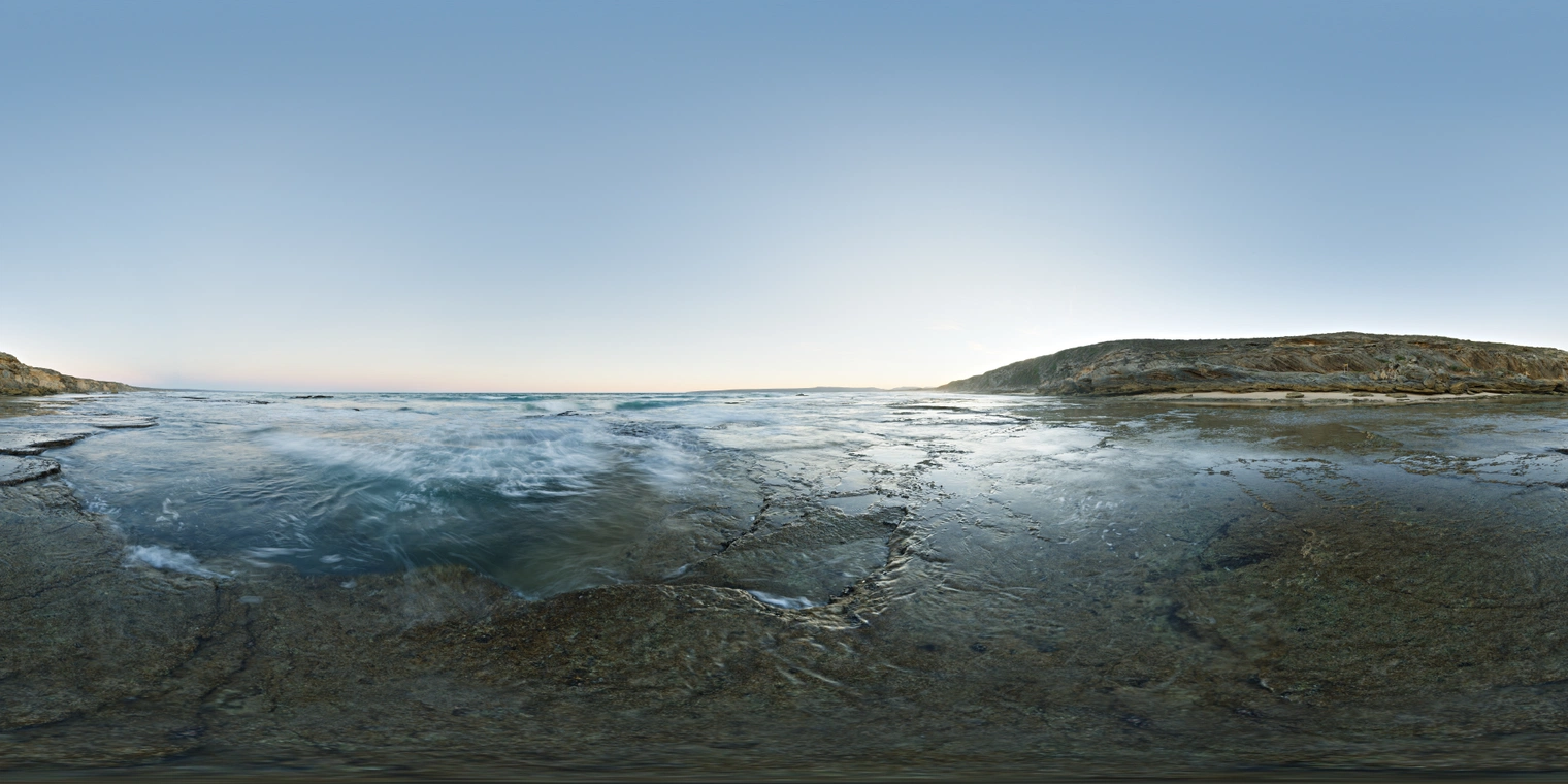 Unclipped, free 24K coastal sunrise HDRI: clear sky, low warm sun over rocky shore and swirling well. Cool blue fill, crisp highlights, soft shadows.