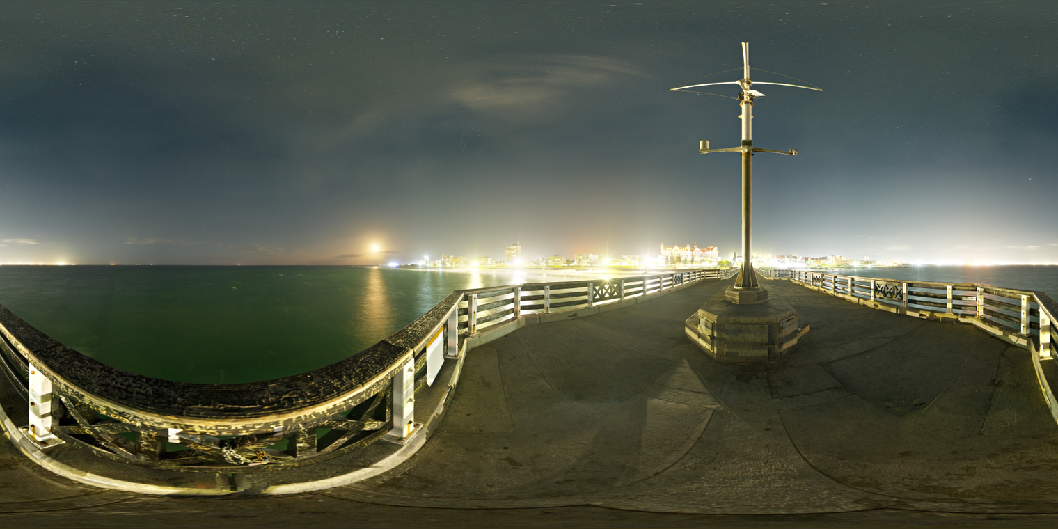 Free, unclipped 24K HDRI of a night pier over the sea, with bright city glow, starry sky, cool teal water, warm lamps, sharp highlights and soft shadows.