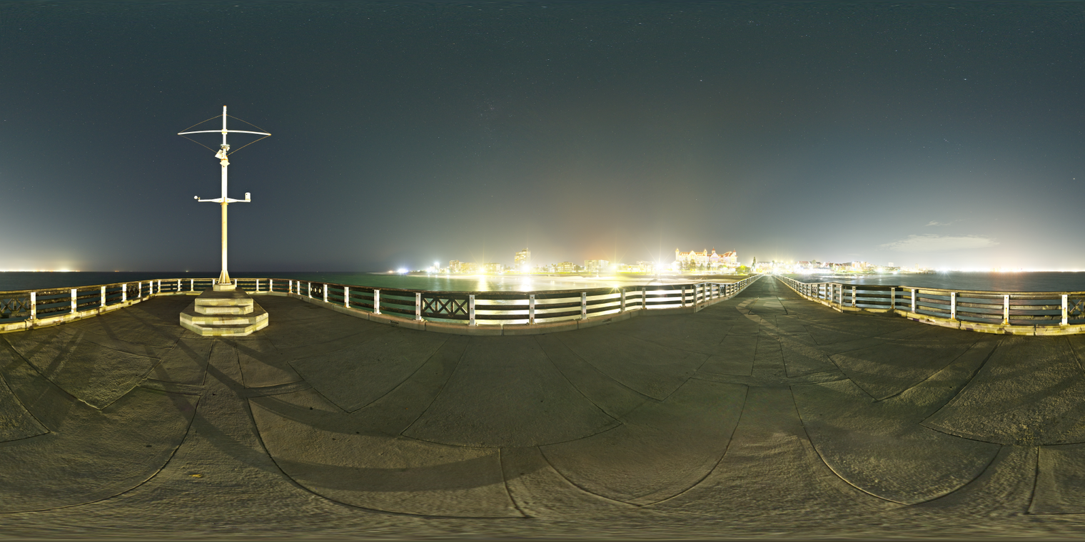 Unclipped, free 24K HDRI of an ocean pier at night: starry sky, dark sea, warm city glow on the horizon, crisp specular lights, concrete pier and railings.