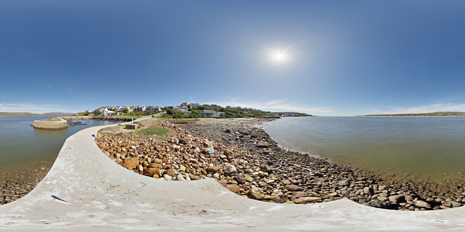 Unclipped, free 24K HDRI of a sunny harbor: clear blue sky, high sun, hard shadows, bright reflections over calm water and a warm, rocky shore.