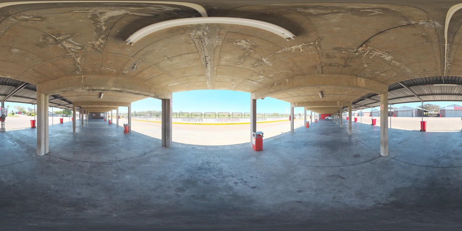 Free, unclipped 20K HDRI. Midday racetrack pit with covered concrete bays: bright sun outside, cool shaded interiors, medium-contrast natural light.